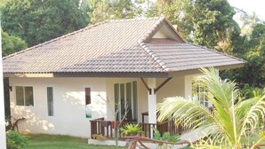 Une maison de plain-pied avec un toit en tuiles, une véranda dotée d’une balustrade et un petit jardin agrémenté de plantes tropicales.