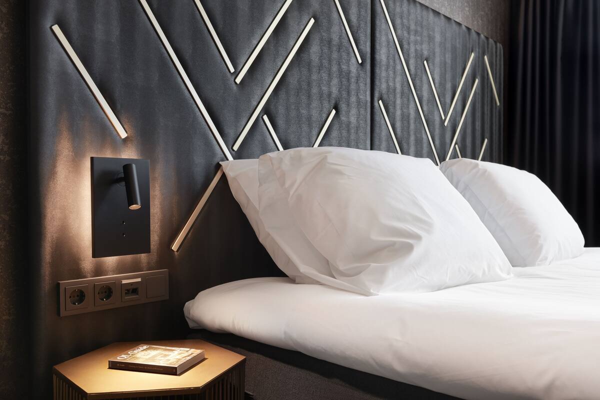 Une chambre d’hôtel moderne avec un grand lit, du linge de lit blanc, une lampe fixée au mur et une table de chevet avec un livre.