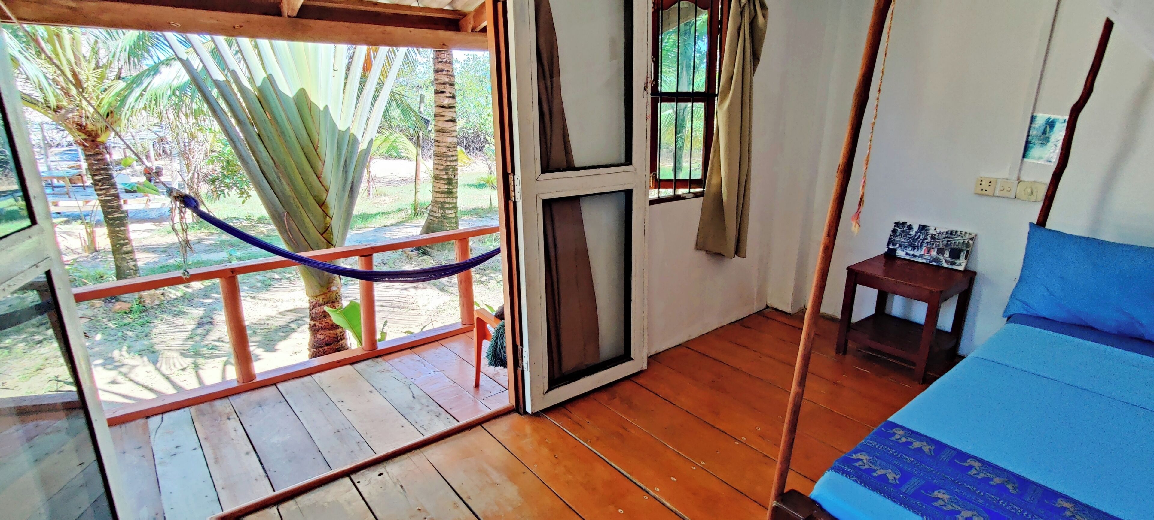 A room with a wooden floor, a bed with a blue bedspread, a small table with a picture frame, a window with a curtain, and a view of palm trees and a car outside.