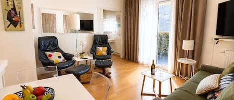 Ein modernes Wohnzimmer mit einer Couch, zwei Sesseln, einem Esstisch mit Früchten, einem Fernseher und einem Balkon mit Vorhängen.