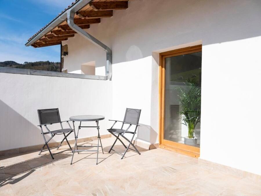 Um pátio externo com mesa e duas cadeiras, uma pérgola de madeira e uma porta de vidro que dá para uma área verde.