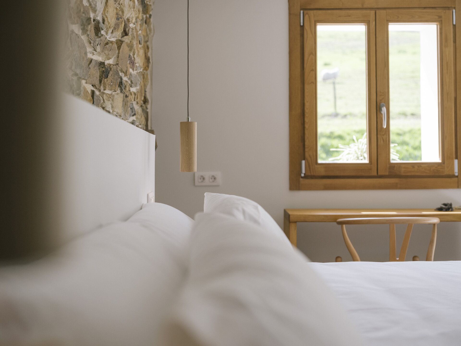 A bedroom with a stone wall, a wooden headboard, a wooden nightstand, and a window with a view of greenery.