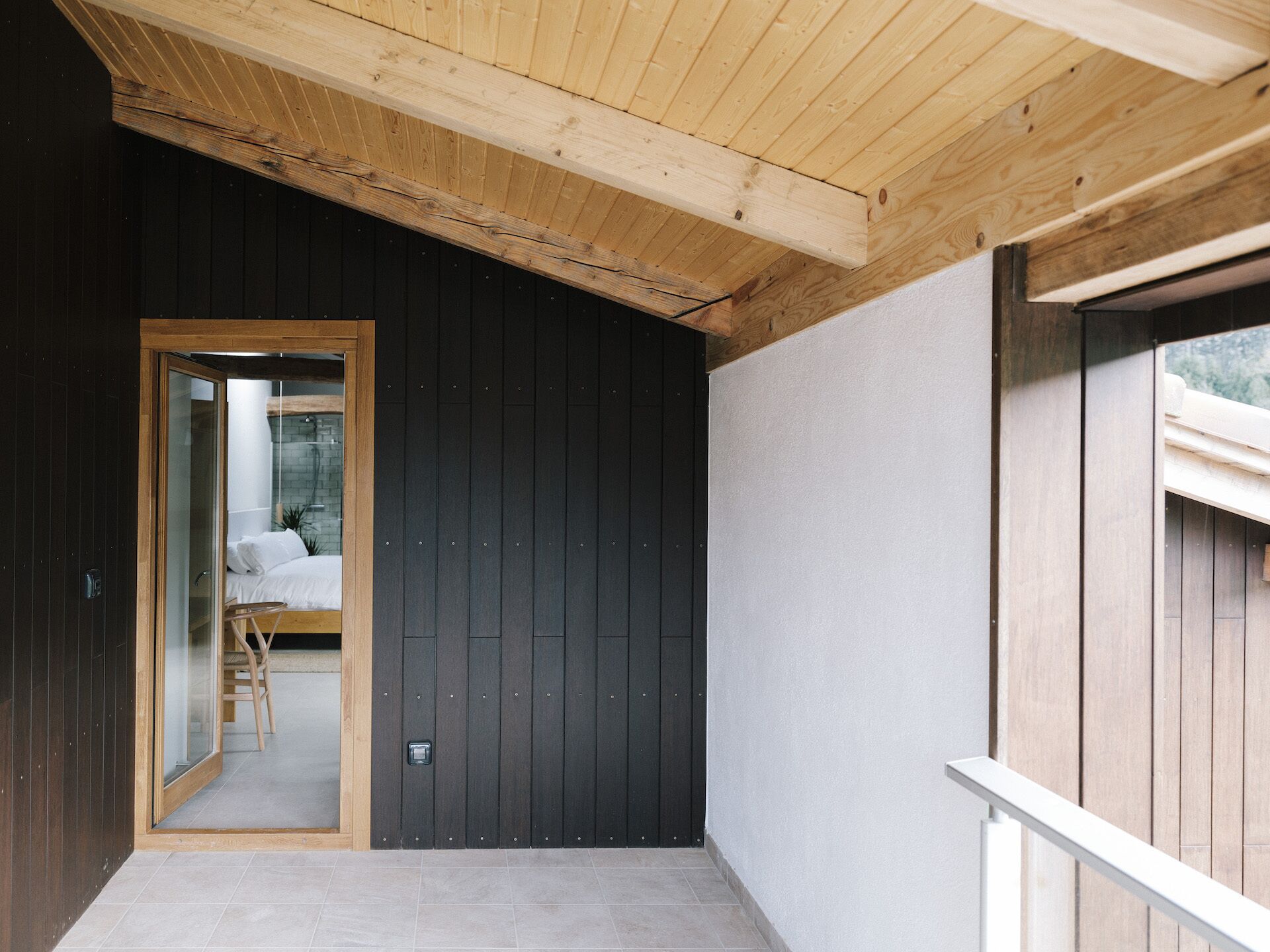 Un interior moderno con una pared de madera oscura, un techo de madera y un vidrio que da a una habitación con una cama y una silla.