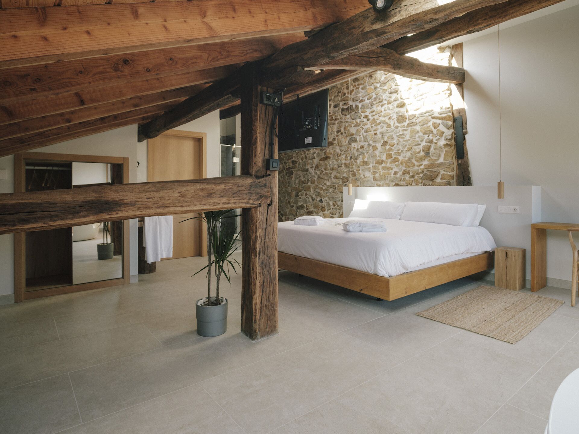 A bedroom with a stone wall, wooden beams, a bed, a nightstand, and a potted plant.
