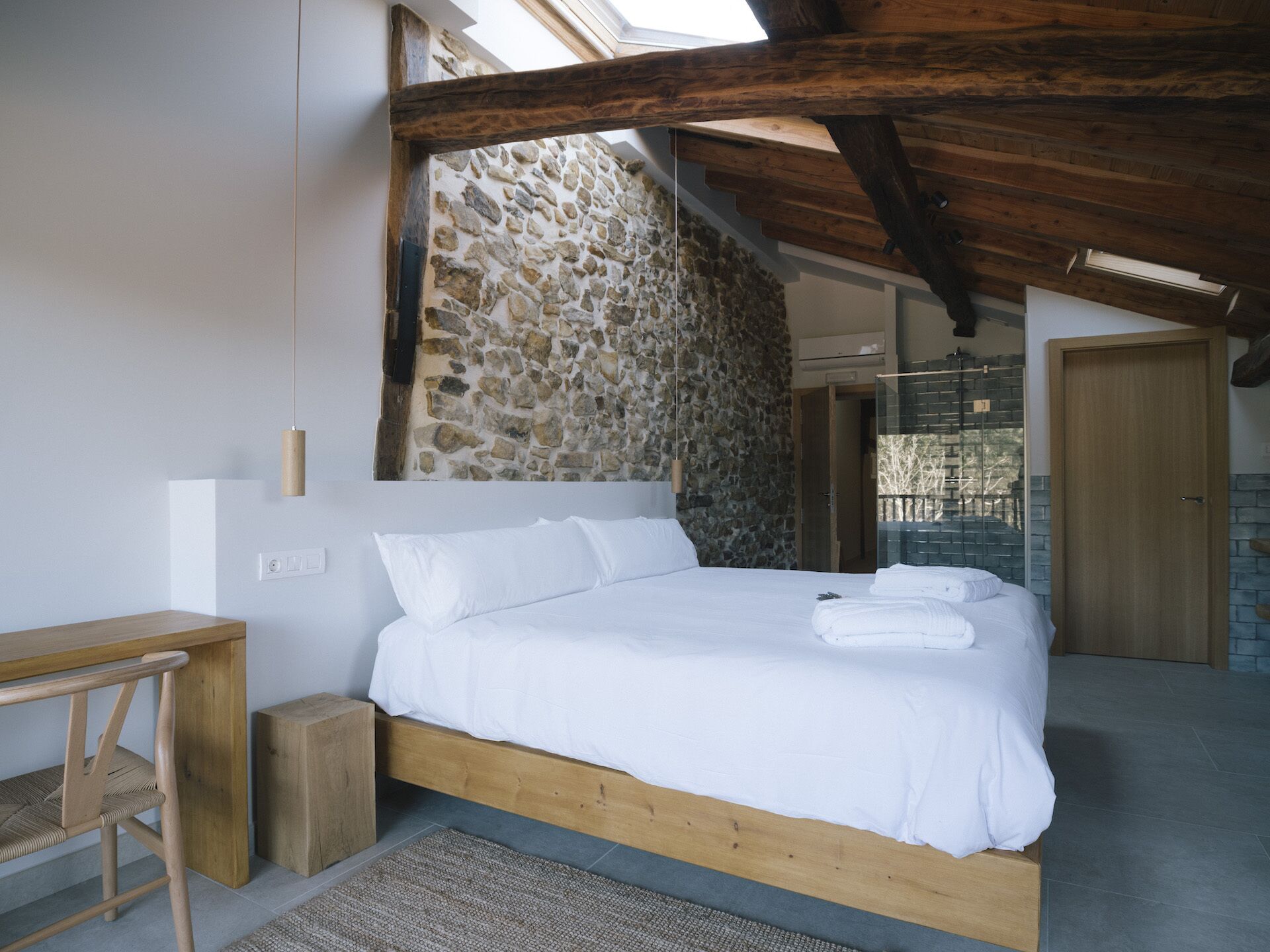A bedroom with a stone wall, wooden beams, a bed with white linens, a wooden desk, and a small rug.