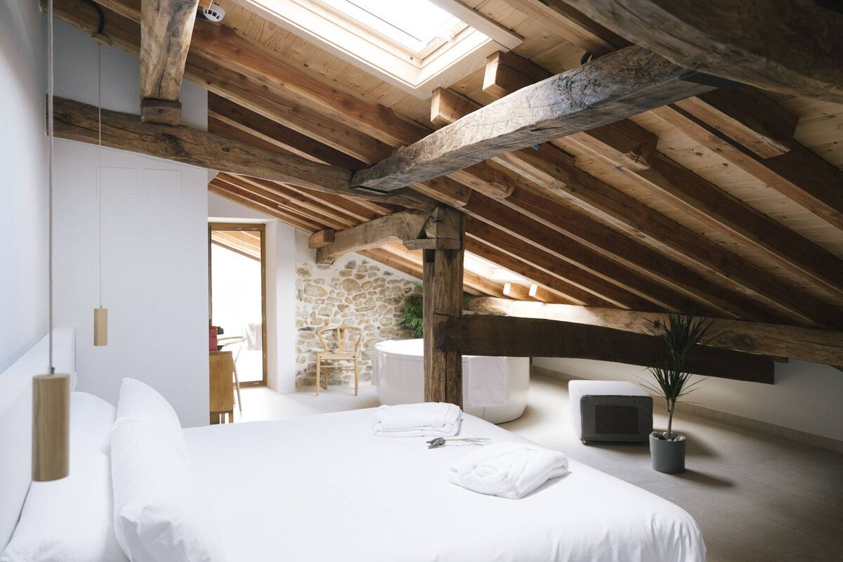 Ein Schlafzimmer mit schrägema Dach, einem Bett mit weißer Bettwäsche, einer Steinwand und einem kleinen Tisch mit Stühlen.