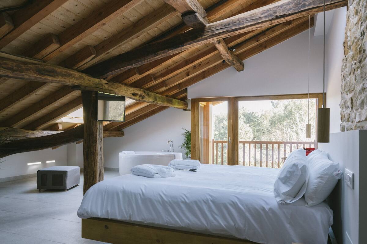 Ein Schlafzimmer mit Holzbalkendecke, einem großen Fenster, einem Bett, einem Nachttisch und einem an der Wand befestigten Spiegel.