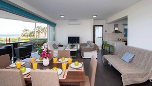 A modern living room with a dining area, a sofa, and a TV. There is a view of the ocean through large windows. - Beachfront apartmen located just a stone's throw away from Ayia Triada beach (Paralimni)