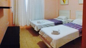 Una camera d'albergo con due letti singoli, un tavolino, una sedia, una finestra con tende bianche e un quadro appeso al muro.
