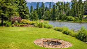 Une zone verdoyante bien entretenue, avec un foyer circulaire en pierre au centre, entourée d’une végétation luxuriante et bordée d’un lac paisible, le tout surmonté d’un paysage montagneux en arrière-plan.