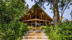 Une cabane en bois avec un toit triangulaire, entourée d’une végétation luxuriante et accessible par un escalier en béton.