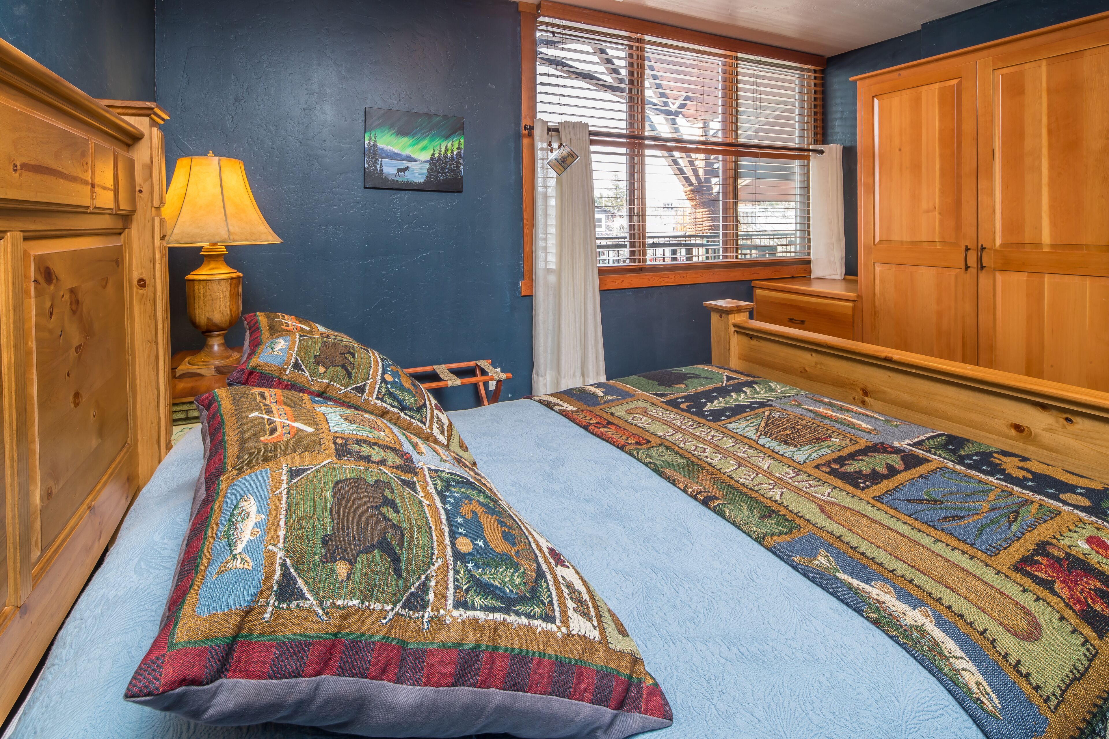 Une chambre à coucher avec un lit, une armoire en bois, une fenêtre avec des stores, une lampe et un tableau au mur.