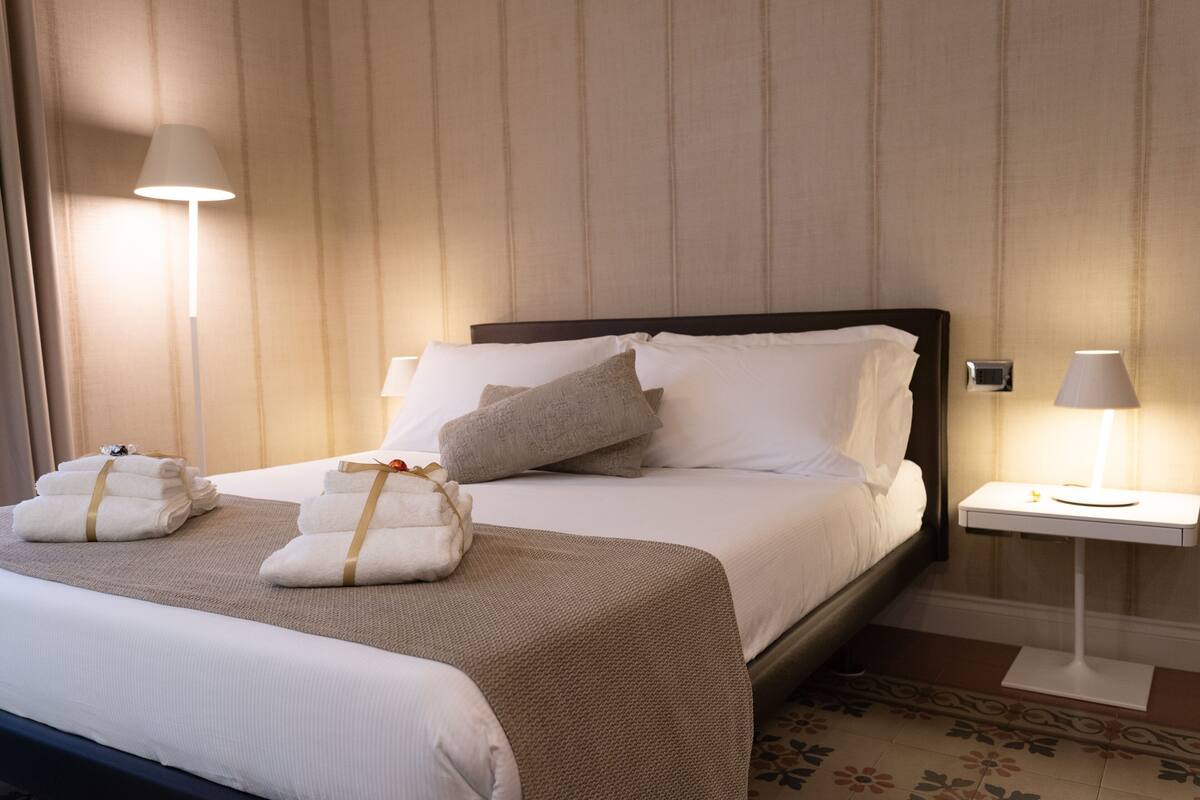 Ein Hotelzimmer mit einem großen Bett, zwei Nachttischen mit Lampen und ordentlich gefalteten Handtüchern auf dem Bett.