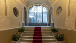 Una imponente escalera con alfombra roja, bordeada por plantas en macetas, que conduce a un amplio ventanal con arco.