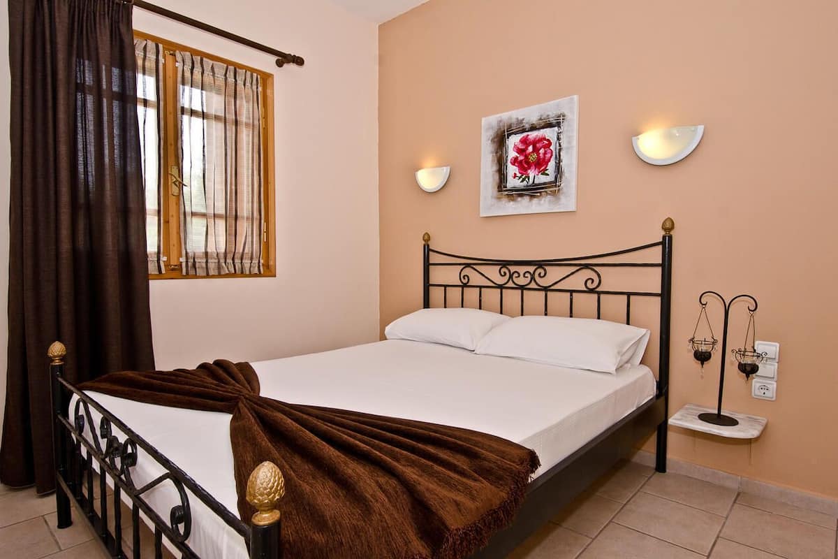 A neatly made bed with a brown blanket, a window with curtains, wall-mounted lights, and a framed picture on the wall.