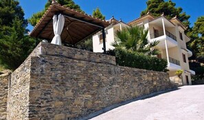 A stone wall with a pergola and a white building with balconies. - Likasti (Skopelos)