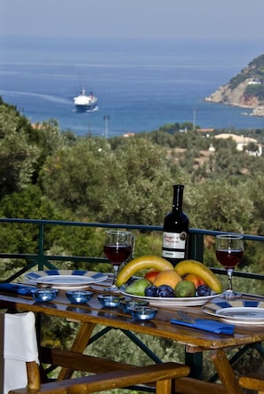 Une table dressée avec une bouteille de vin, des verres et un plateau de fruits, offrant une vue sur la côte et un bateau au loin.