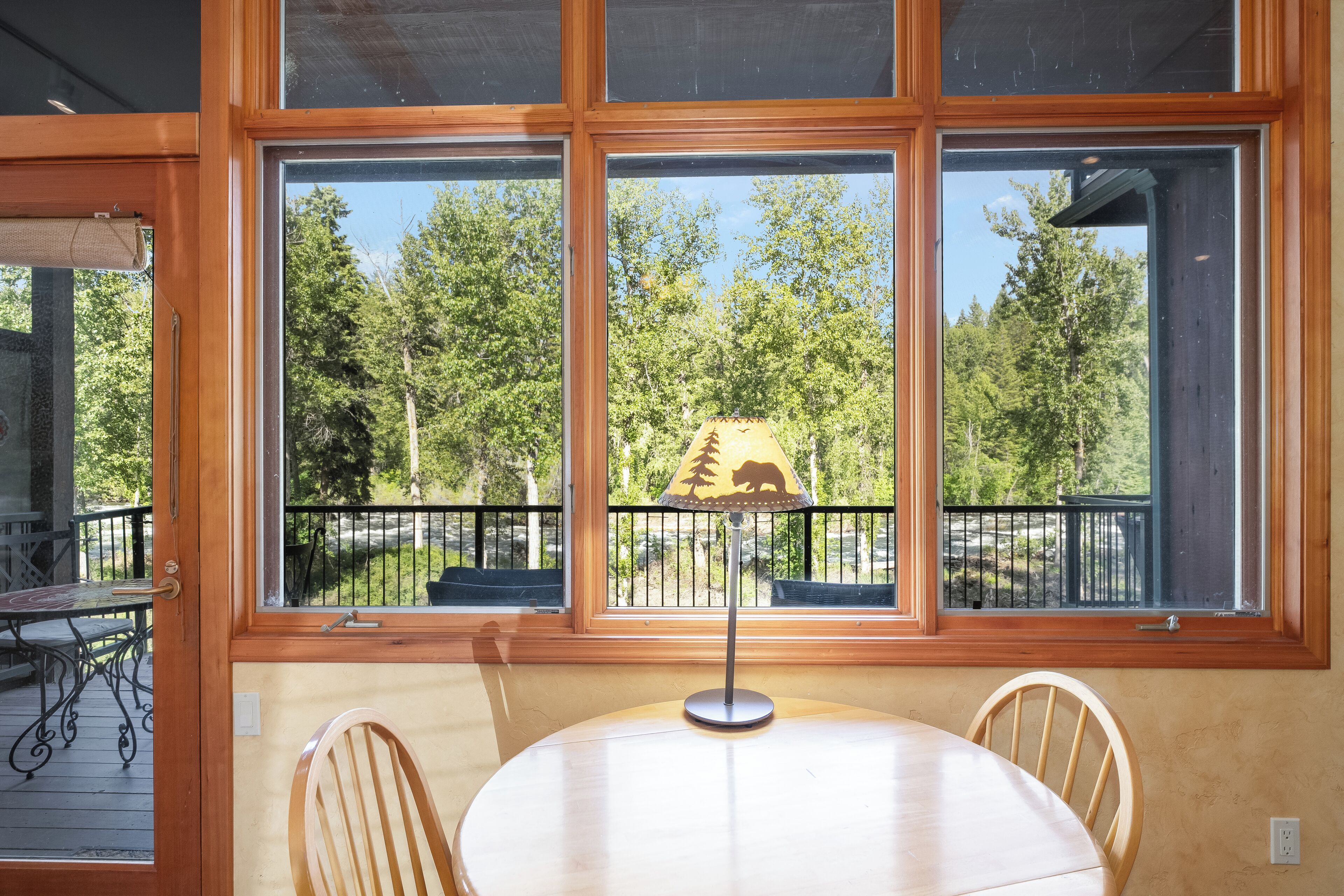 A room with a round table, two chairs, a lamp, and a view of a forest through large windows.