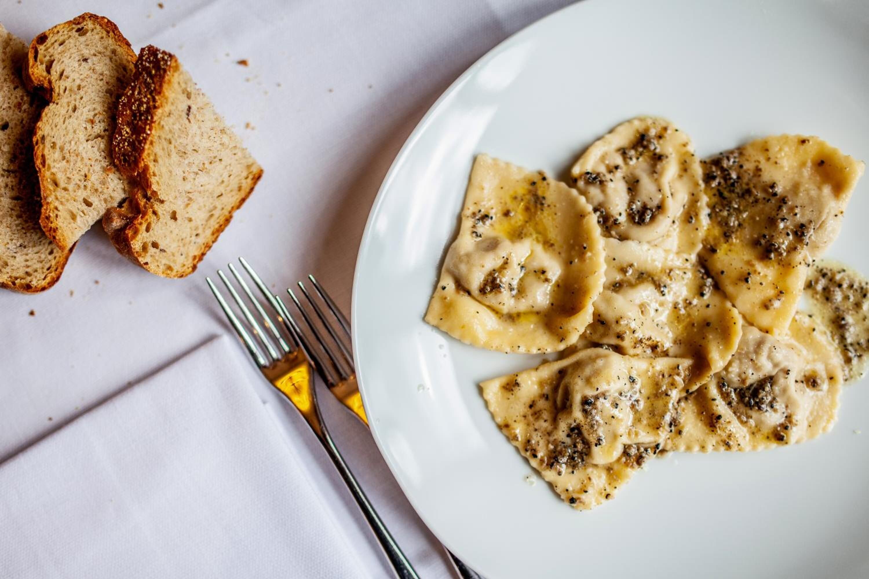 A plate of ravioli with a creamy sauce and black pepper, accompanied by slices of bread on a white plate.