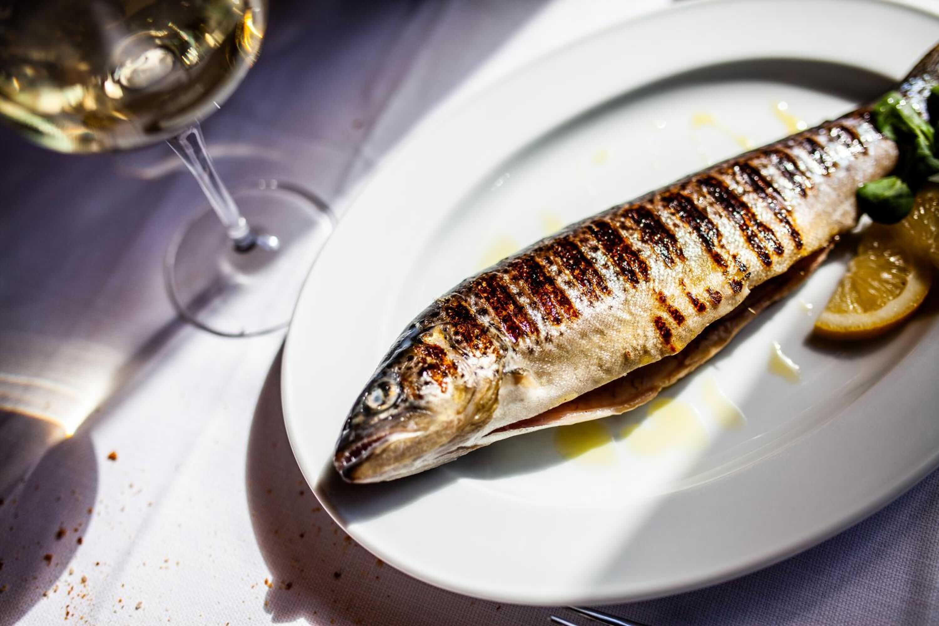 Pescado a la plancha en un plato con rodajas de limón y una copa de vino blanco.