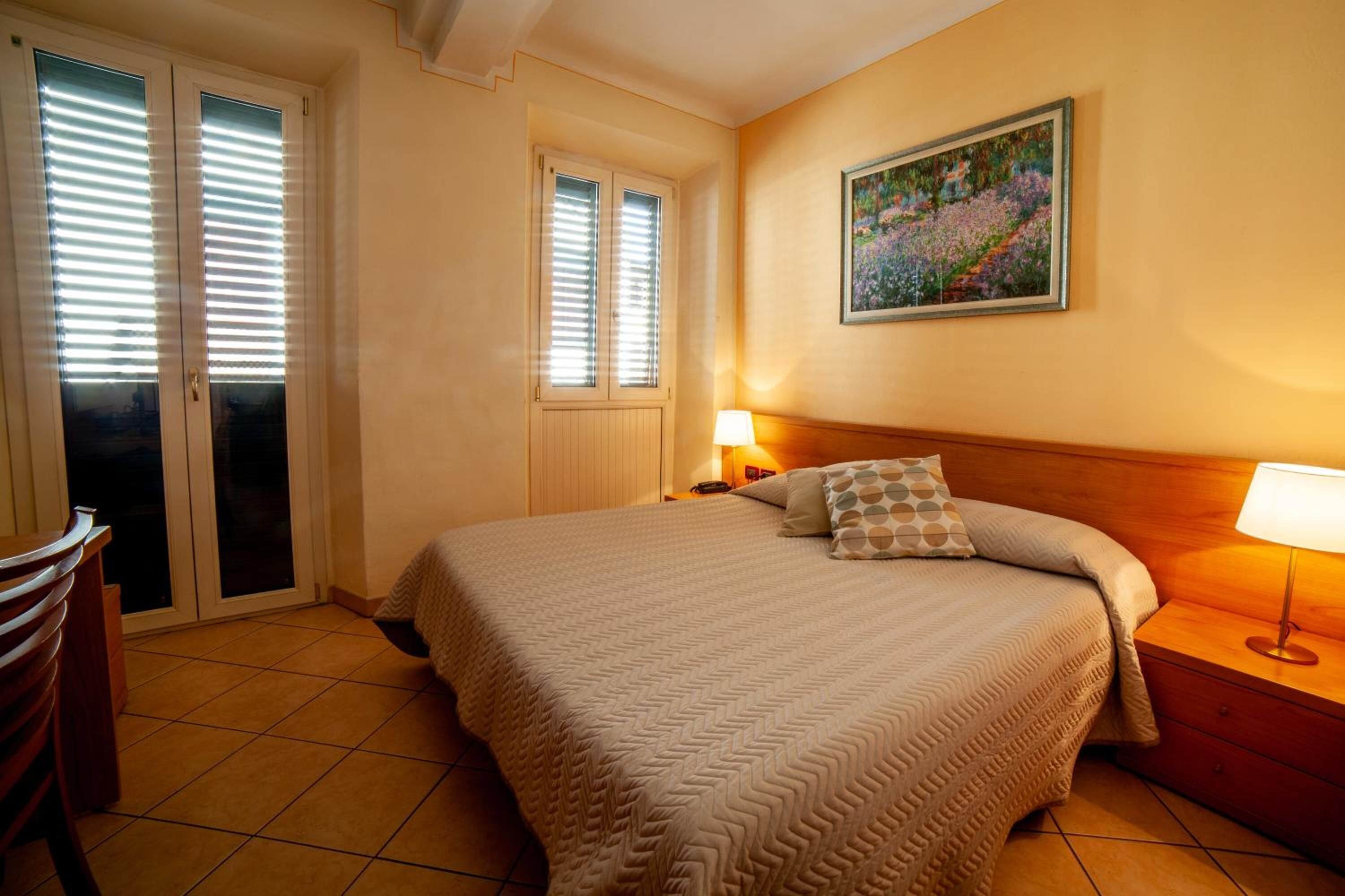 Een slaapkamer met een groot bed, een nachtkastje met een lamp, een schilderij aan de muur en een deur met jaloezieën.