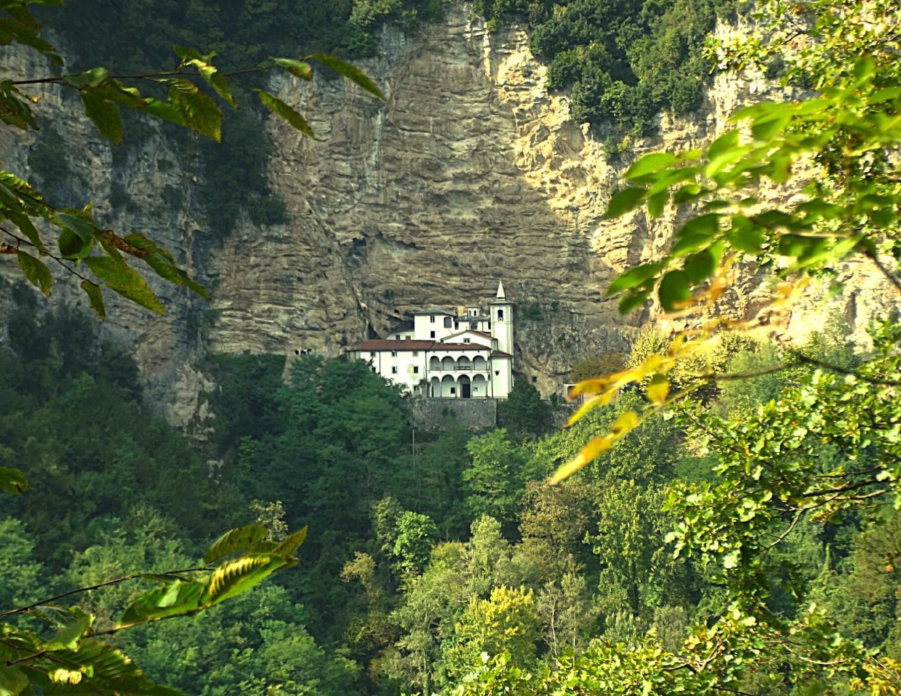 Un edificio blanco con un campanario, ubicado en un acantilado rocoso rodeado de un bosque denso.