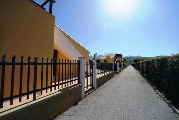 A pathway flanked by a black metal fence and a yellow building with a sloped roof.
