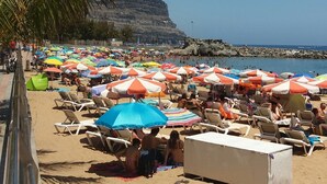 A beach with rows of sun loungers and umbrellas, a palm tree, and a rocky cliff in the background.