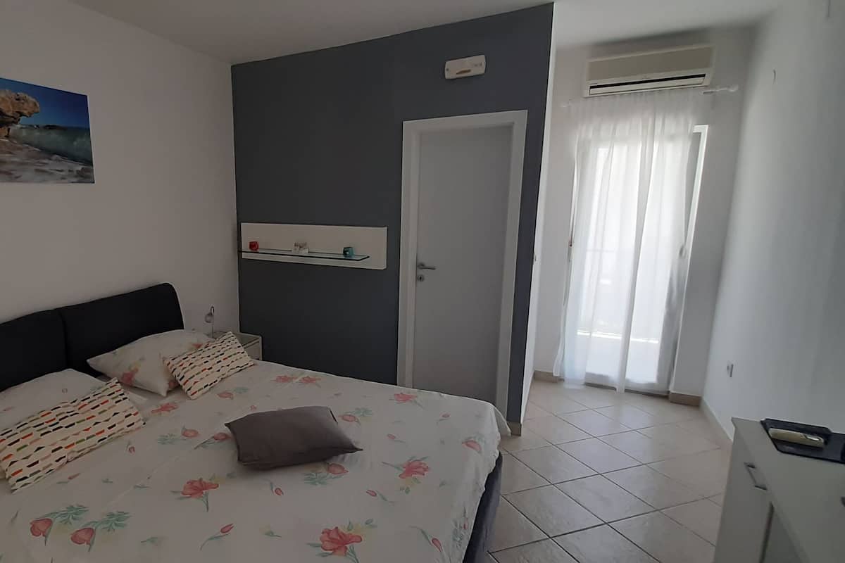 A bedroom with a bed, a nightstand, a wall-mounted shelf, a door, and a window with curtains.
