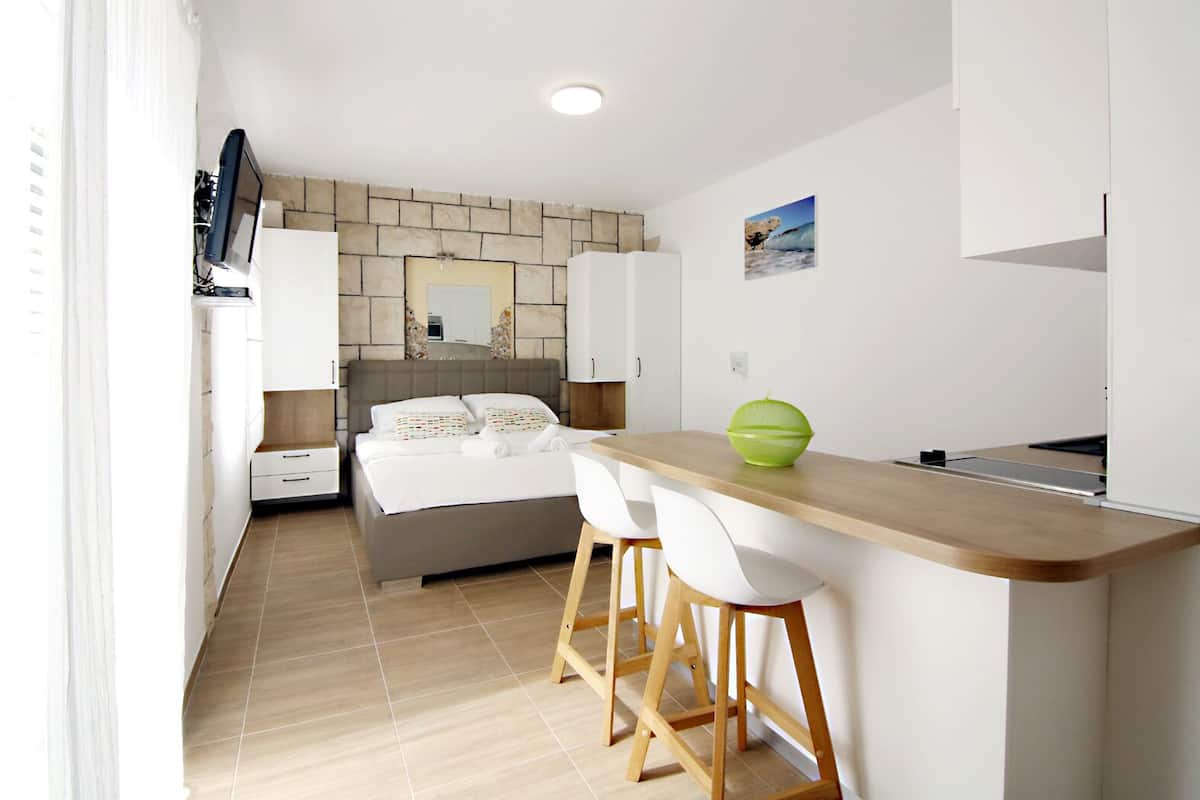A small, modern bedroom with a bed, a nightstand, and a desk with two chairs. There is a window on the left side.