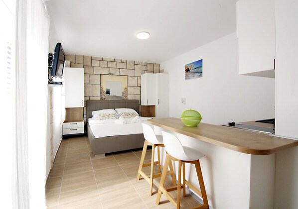 A small, modern bedroom with a bed, a nightstand, and a desk with two chairs. There is a window on the left side.