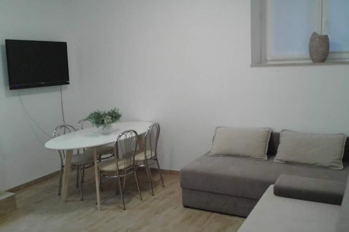 A living room with a grey sofa, a white dining table with metal chairs, and a flat-screen TV mounted on the wall.