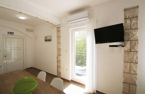 A room with a dining table, chairs, a green bag, a TV mounted on a stone wall, and a door with an emergency exit sign.
