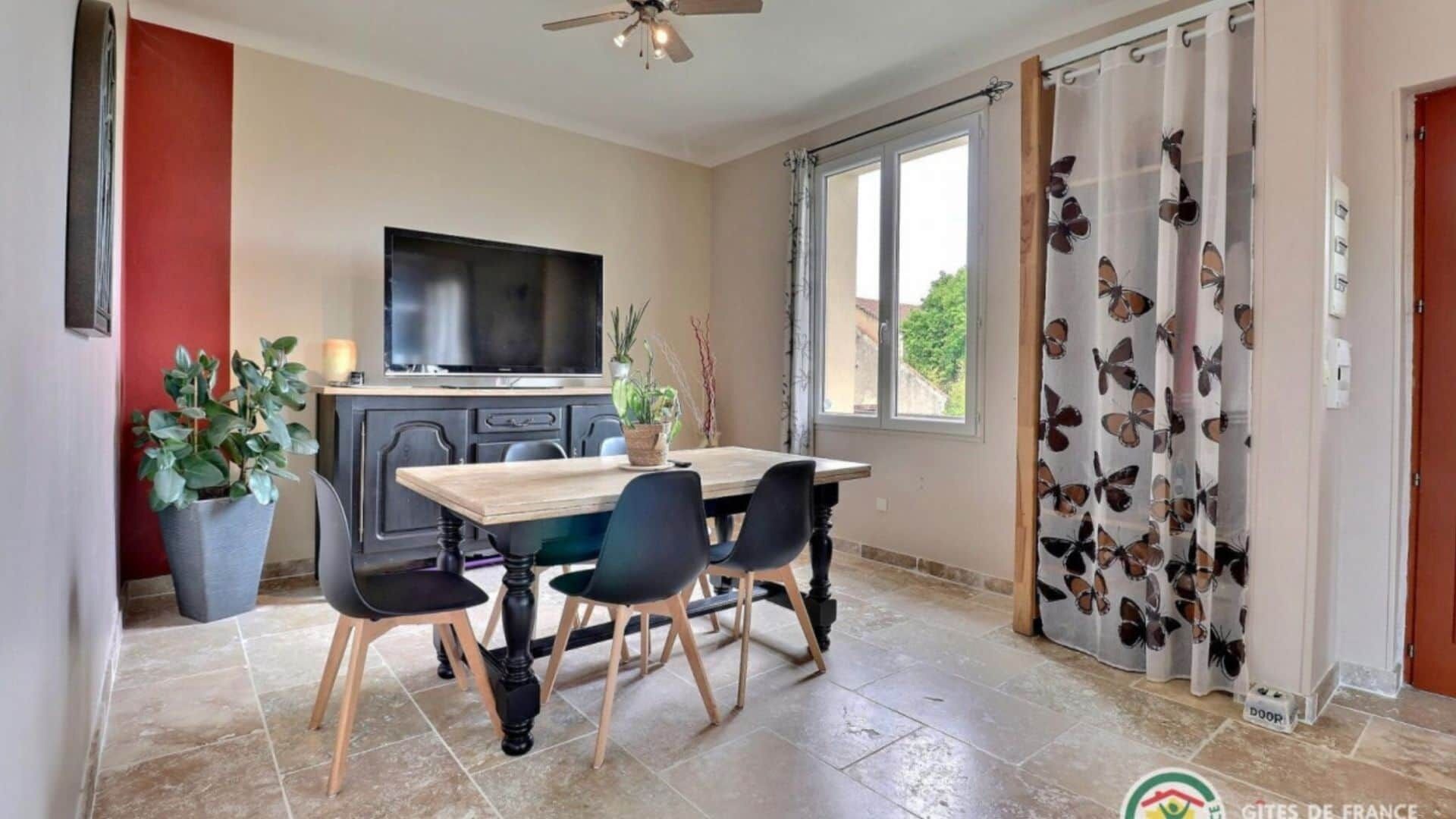 Uma sala de jantar com mesa e cadeiras de madeira, televisão sobre um móvel e janela com cortinas.