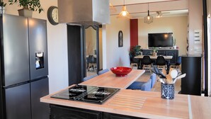A modern kitchen with a built-in stove, a red bowl on the counter, and a dining area with a TV and seating. - Les Gîtes de L'Aven - la Forestiere (Orgnac L'aven)