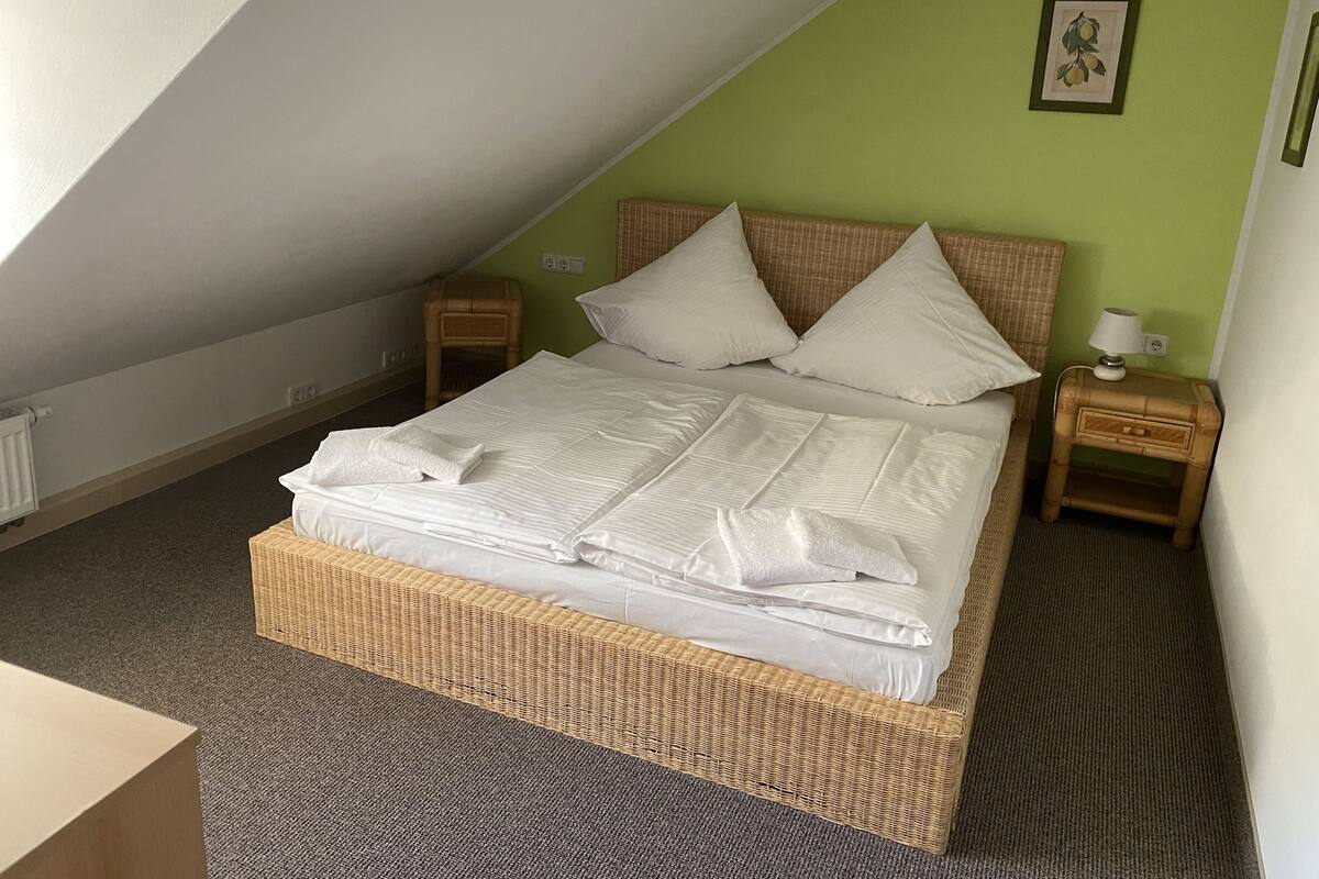 Una cama bien tendida en una habitación con techo inclinado, una mesita de noche con lámpara y un cuadro enmarcado en la pared.