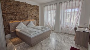 A modern bedroom with a stone accent wall, a large bed with white linens, a wooden headboard, a marble floor, a vanity with a mirror, and sheer curtains with a striped pattern. - Hotel Sächsischer Hof (Scheibenberg)