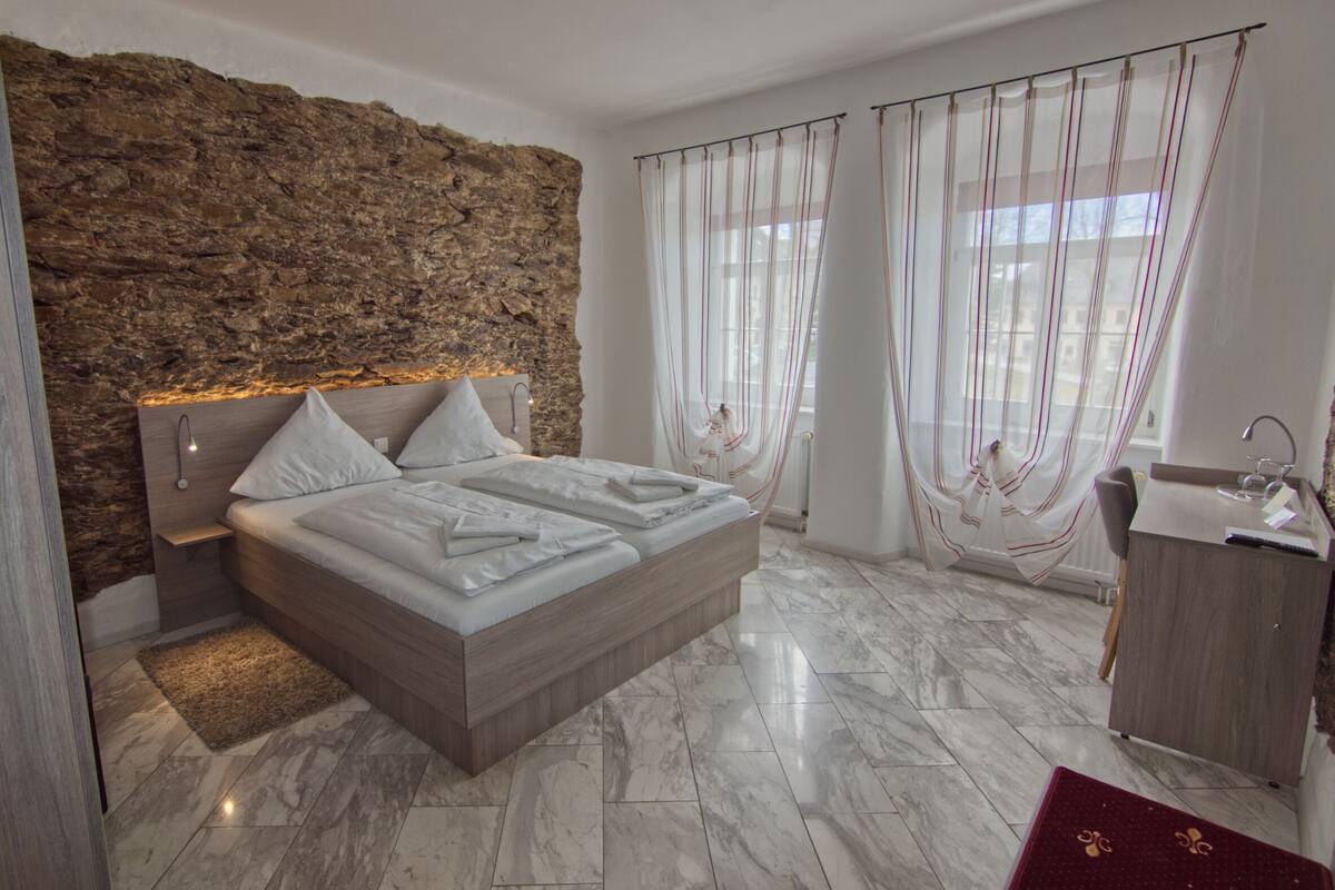 Un dormitorio moderno con una pared de acento de piedra, una cama grande con ropa de cama blanca, un cabecero de madera, suelo de mármol, una tocador con espejo y cortinas de gasa.