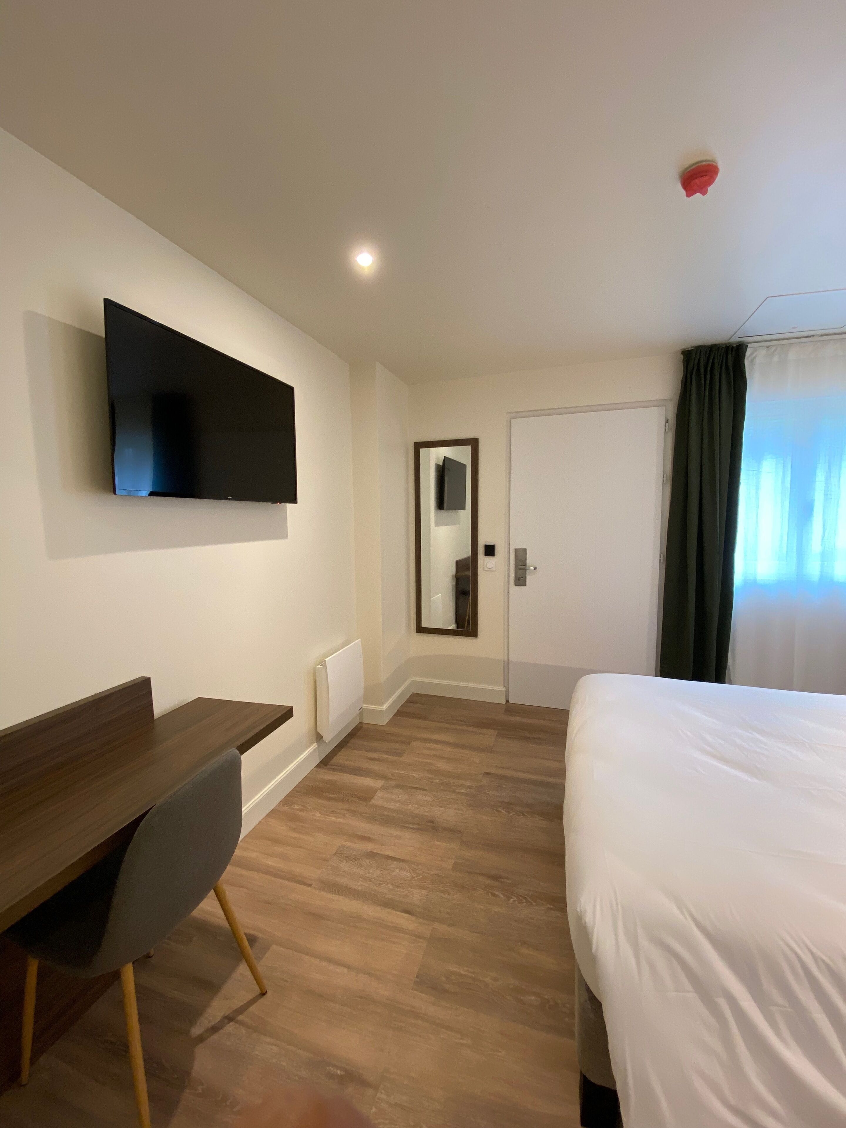 Een hotelkamer met een bed, een bureau, een stoel, een aan de muur bevestigde televisie, een deur en een raam met gordijnen.