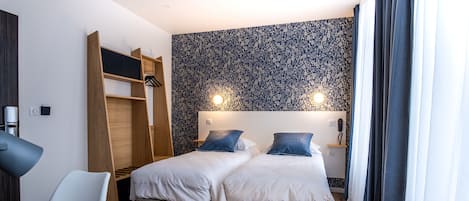 A hotel room with two beds, a desk, a chair, a wardrobe, and a patterned wall.