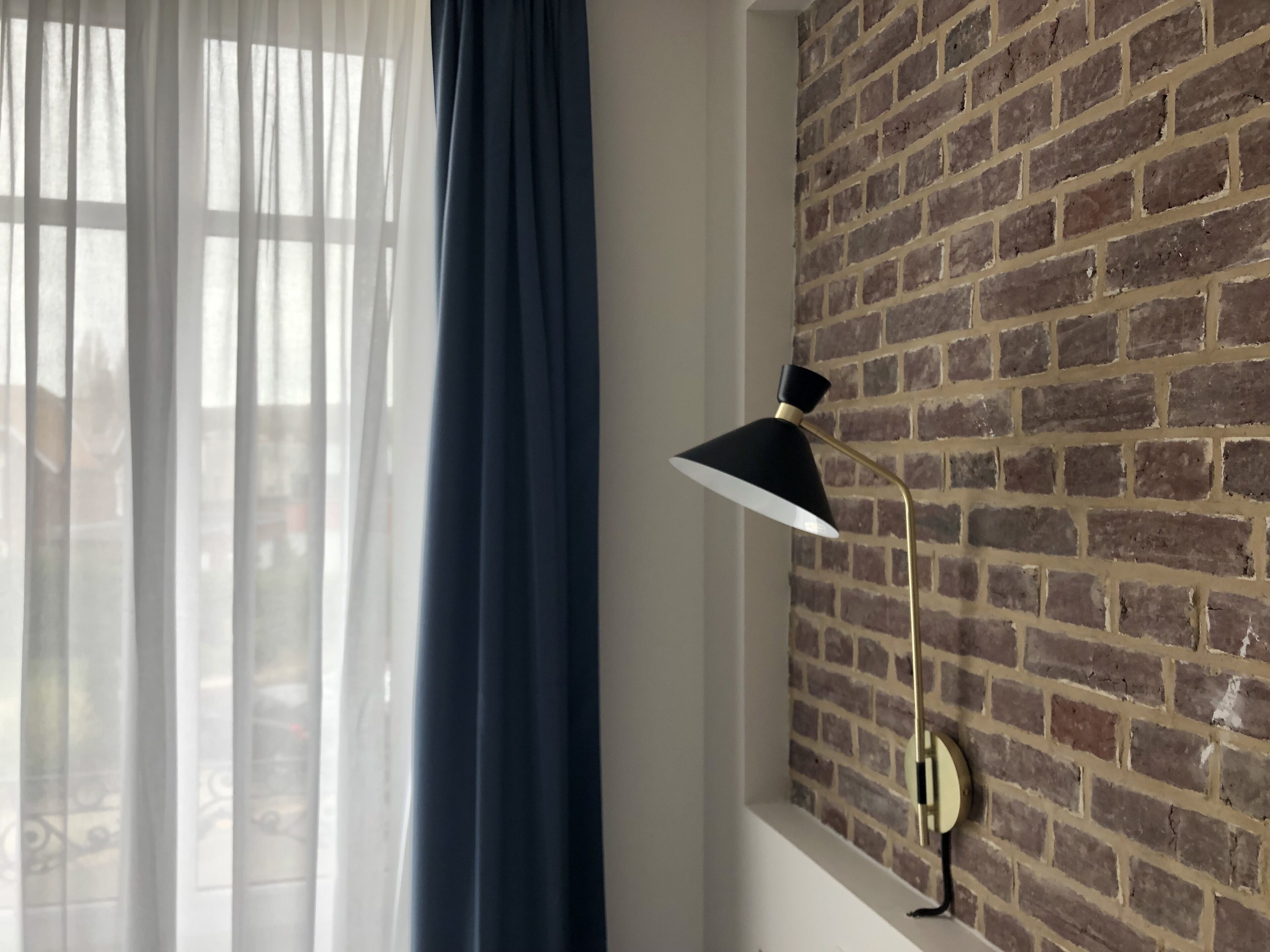 Una habitación con pared de ladrillo, una ventana con cortinas translúcidas y una lámpara fijada a la pared.
