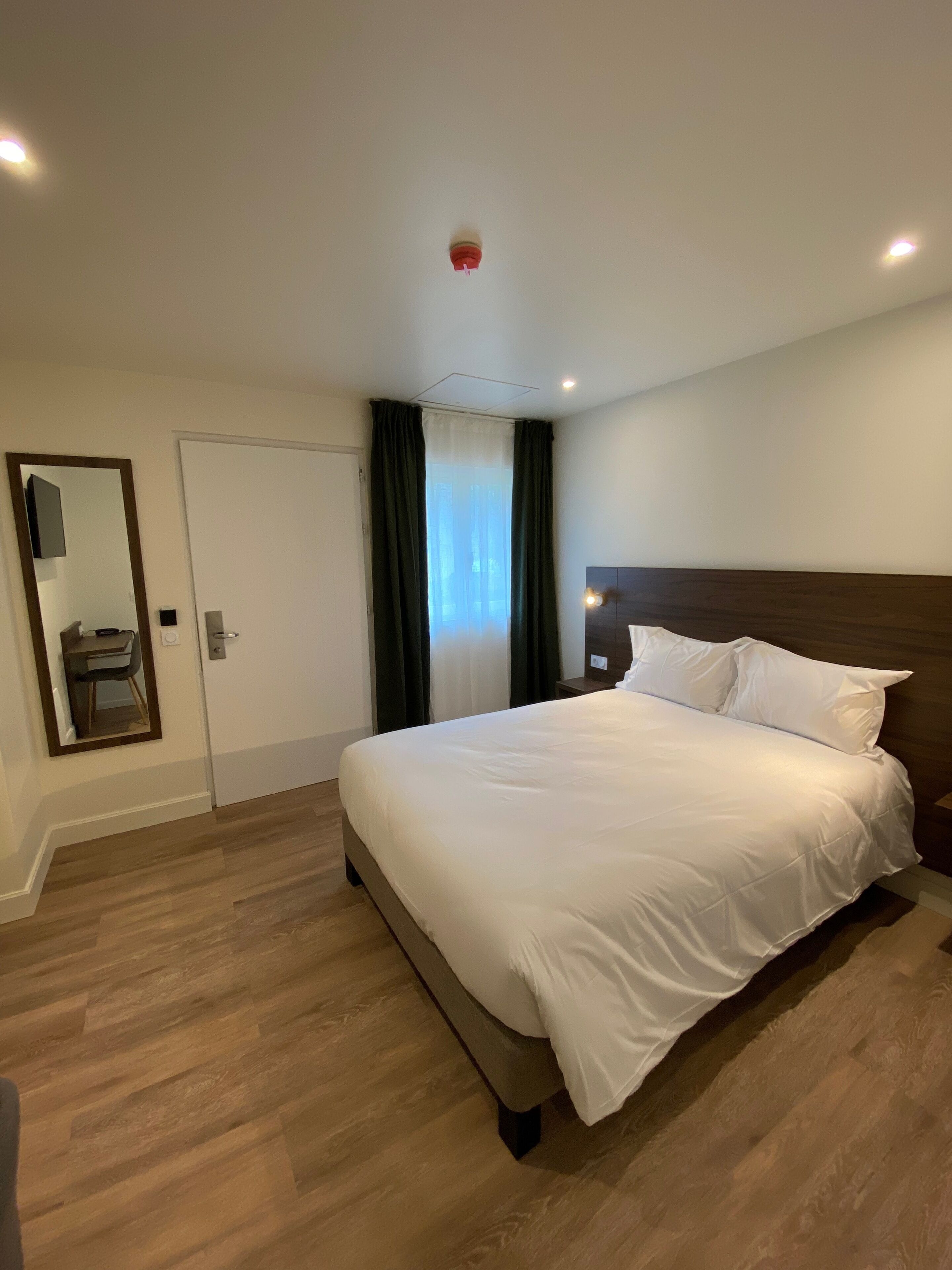 Een hotelkamer met een groot bed, nachtkastje, spiegel, deur en raam met gordijnen.