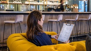 Una persona sentada en una silla amarilla en una zona de bar, sosteniendo un portapapeles con una lista.
