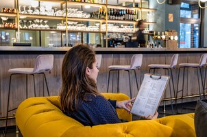 Una persona sentada en una silla amarilla en una zona de bar, sosteniendo un portapapeles con una lista.