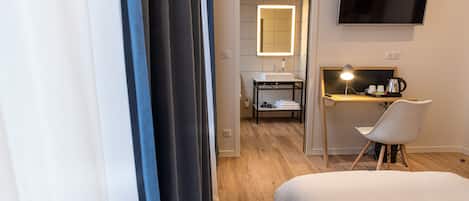 A hotel room with a bed, a desk with a chair, a TV mounted on the wall, and a bathroom with a mirror and sink.