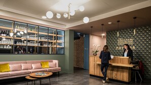 Un área de bar moderna con un mostrador de recepción de madera, un sofá rosa con cojines amarillos y un amplio ventanal que muestra diferentes botellas y vasos.