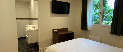 A hotel room with a bed, a TV mounted on the wall, a window with curtains, a shower area, and a small bedside table.