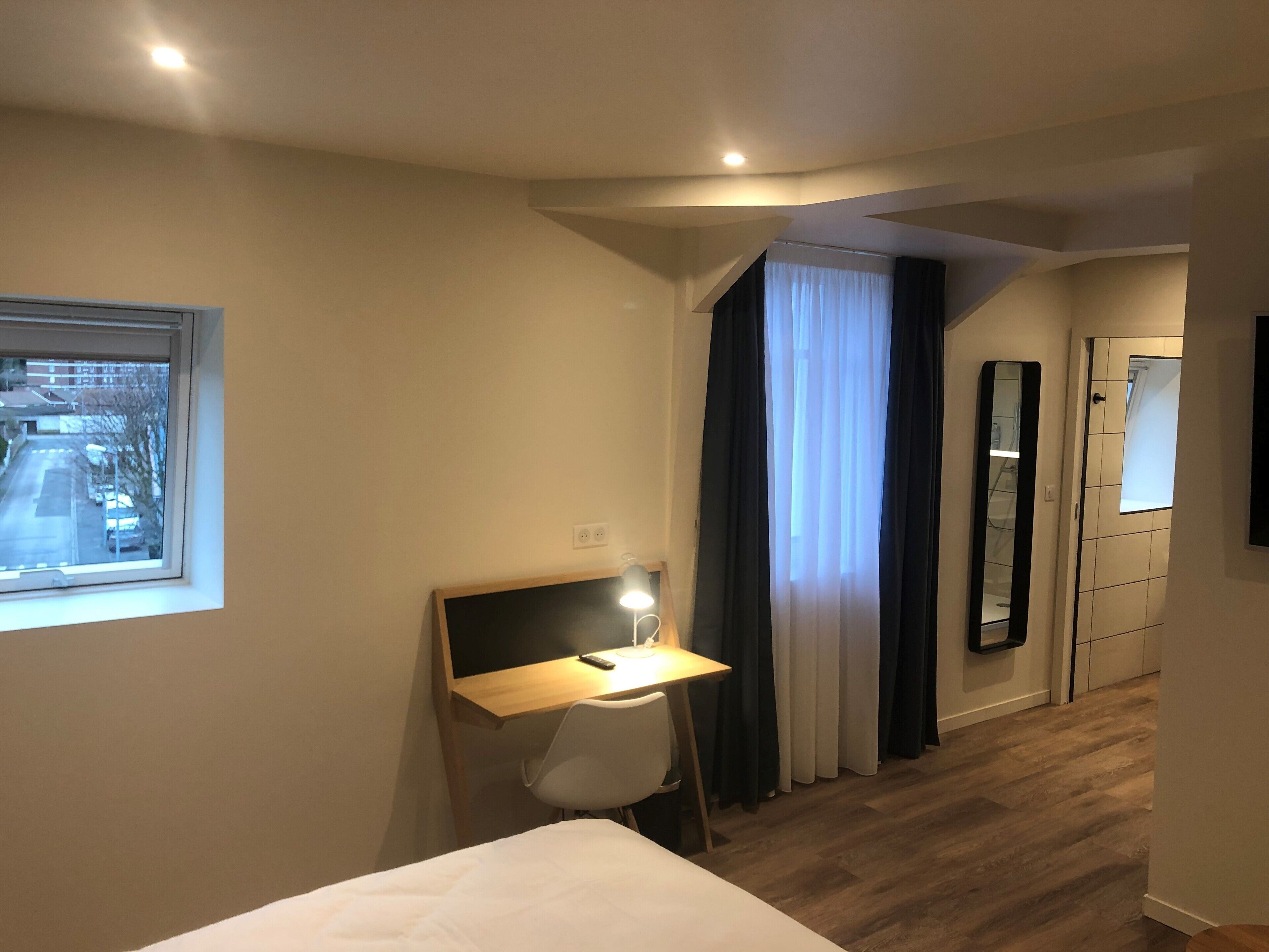 A hotel room with a bed, a desk with a lamp, a window with a view of the street, and a door leading to a bathroom.
