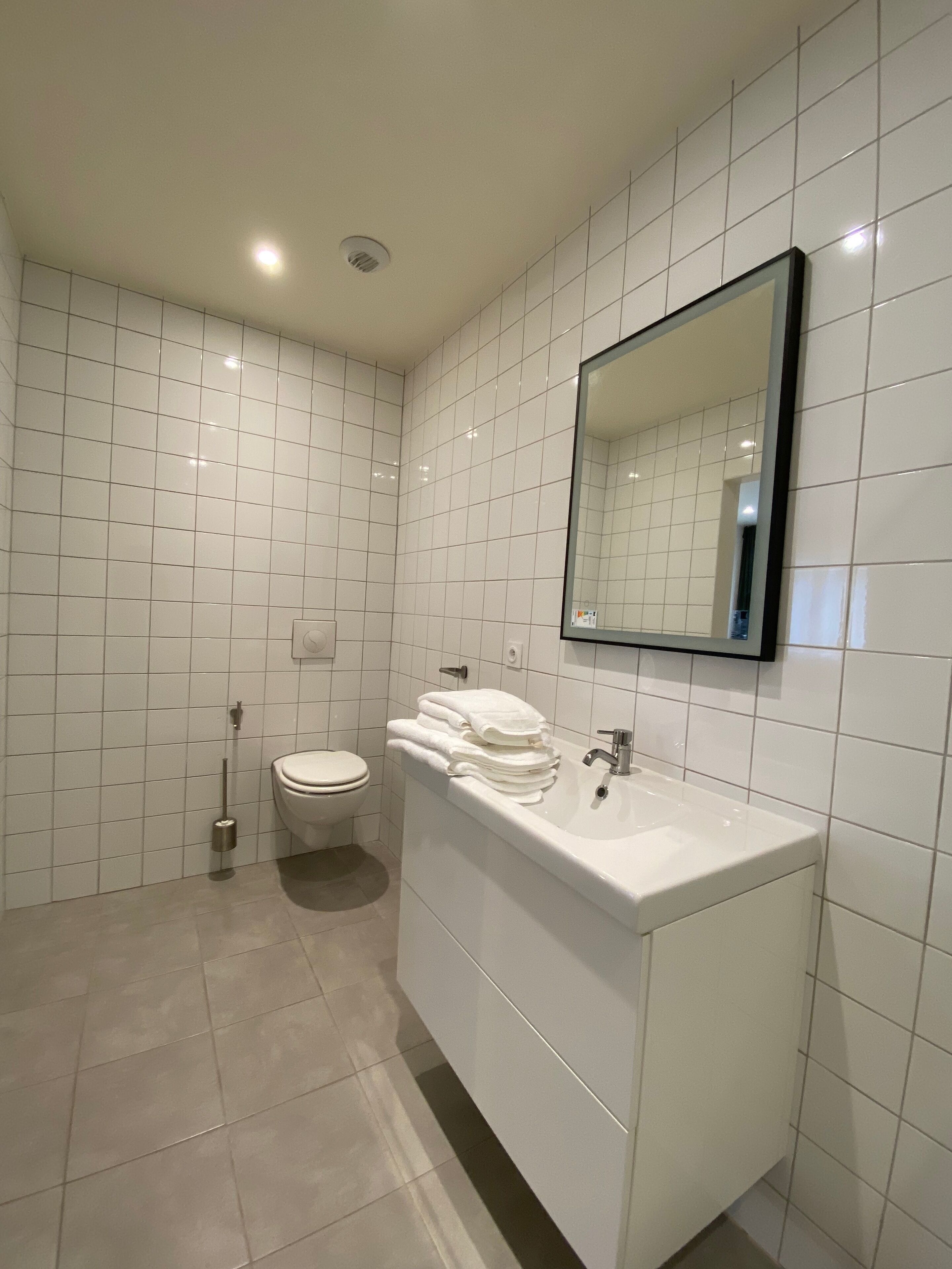 Une salle de bain moderne avec des carreaux blancs, des toilettes, un lavabo et un miroir.