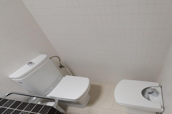 Un baño pequeño y limpio con inodoro, bidé, paredes de azulejos y un lavabo de metal.